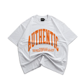 A white oversized short-sleeve t-shirt with a bold orange graphic reading 'AUTHENTIC' and smaller text below, laid flat on a gray concrete surface.